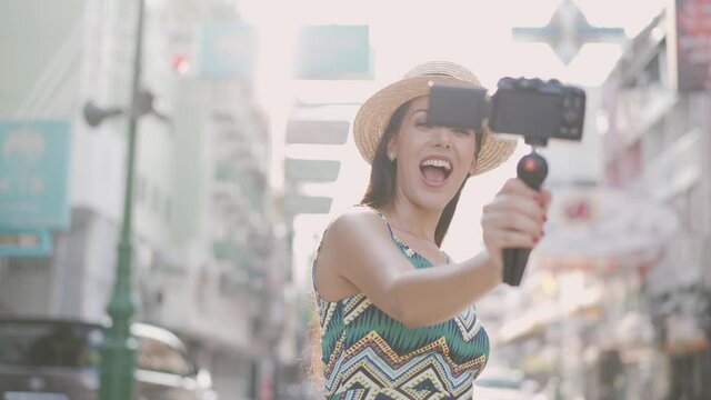 Young brunette woman using a video camera to record journey with smile. Happy and excited 20s Hispanic girl traveler wear summer dress with hat waving to internet audience. Influencer and vlogging