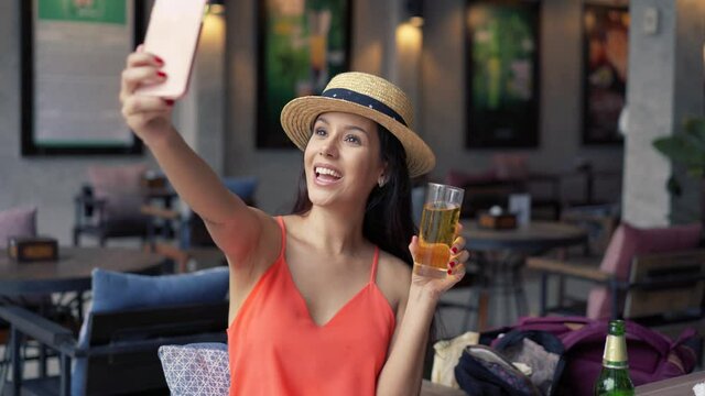 Portrait of young brunette woman taking a selfie photo from cell phone in pub bar. 20s Hispanic girl in summer hat and clothes using mobile phone to video call people in indoors