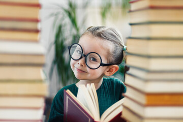 Cute smiling preschool age girl in round big glasses with red book looking at a camera between two books mass