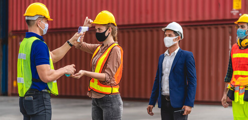 Professional of group cargo foreman in helmets standing and using infrared thermometer for checking body temperature staff fever before work in quarantine for coronavirus wearing protective mask