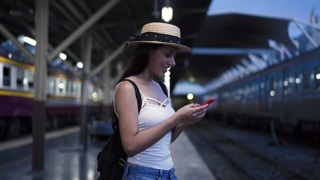 Young brunette woman traveler laughs while playing a mobile phone in train station platform. 20s Hispanic girl passenger in summer clothes on vacation trip. Transportation and technology concept