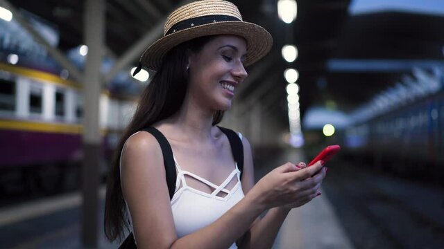Young brunette woman traveler laughs while playing a mobile phone in train station platform. 20s Hispanic girl passenger in summer clothes on vacation trip. Transportation and technology concept