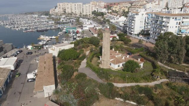 The Punta Doncella lighthouse stands at the port of Estepona. In the background are the boat marina and the houses of the city. The lighthouse is surrounded by trees.