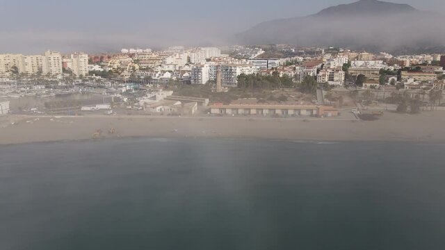 The coast and the beach of the city of Estepona in Andalusia in the morning in the fog. In front of it is the lighthouse Punta Doncella. There is fog and haze over the sea and behind the city.