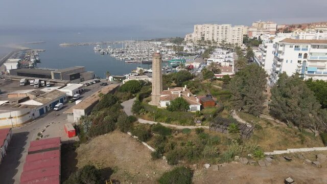 The Punta Doncella lighthouse stands at the port of Estepona. In the background is the boat marina. There is fog and haze over the sea and behind the city.