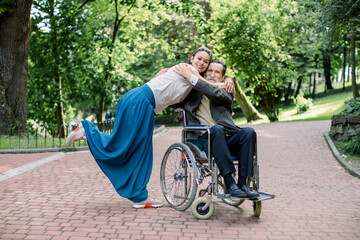 Caring nurse or social worker, pretty young hipster girl walking in park with senior man patient sitting on wheelchair, hugging each other and enjoying summer sunny day.