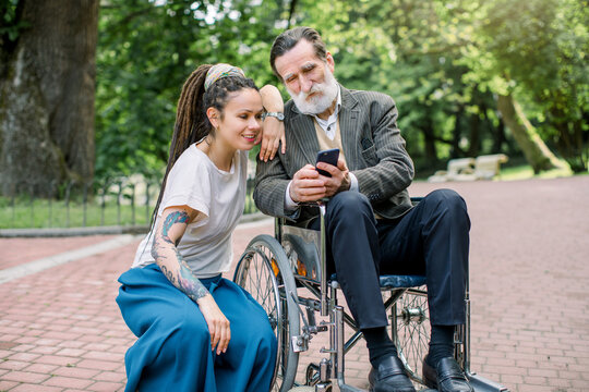 Portrait of happy hipster granddaughter with dreadlocks and barded grandfather in a wheelchair, having fun, making photos or navigating in internet in phone in a green park on a sunny day.