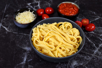 'Tagliolini' noodles, a long ribbon pasta similar to spaghetti in bowl next to small bowl with tomatoe sauce and grated cheese ond dark background