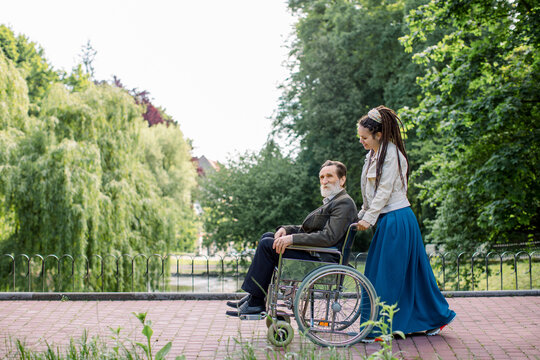 Side view of young modern hipster woman with long dreadlocks, assisting her disabled grandfather on wheelchair in park. Happy elderly man in wheelchair with young girl social worker in the park.