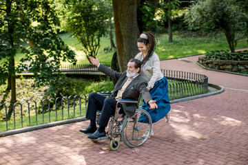 Care helper woman, pretty young hipster granddaughter walking with happy elderly disabled man with arms outstretched, pushing a wheelchair and running in the city park in summer day.