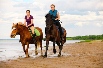 Two happy horsewomen are riding along the beach near the water.