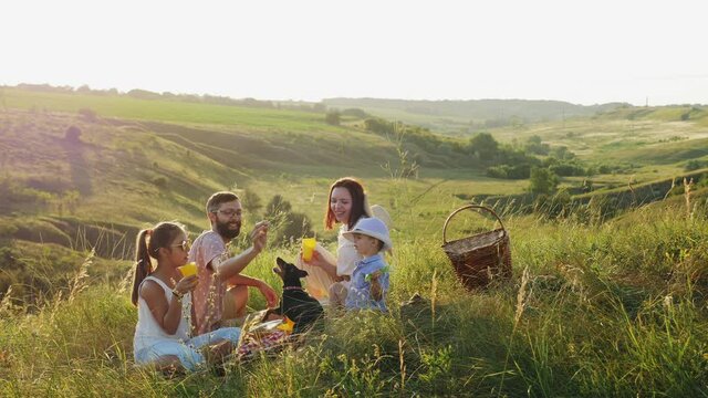 A friendly family of four (with children and a dog) on a picnic in nature. A man feeds a dwarf pinscher from his hand