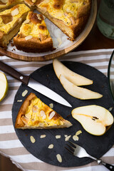 Slice of homemade pear pie and sliced fresh pear on black plate with whole pie and pears on stripped tablecloth .