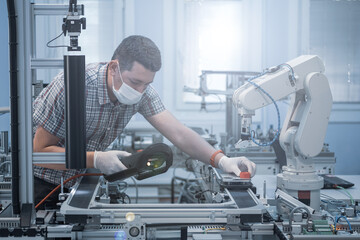 Man with surgical mask and gloves against COVID-19 ( Coronavirus ) programming robot arm with teach pendant. industry 4.0 : smart factory production line control with automation.