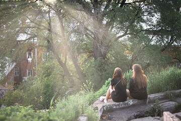 Two young women are sitting on a hillside admiring nature and chatting in the sunray at sunset