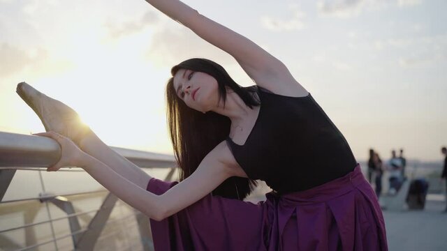 Middle shot of ballerina standing in split and bending aside to leg. Portrait of charming young brunette Caucasian woman performing classical ballet movement on bridge at sunrise.