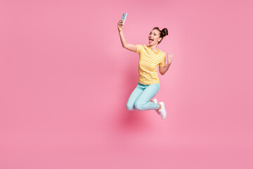 Fototapeta na wymiar Full body photo of attractive lady two buns hold telephone jump up stick tongue making selfies finger horns rocker wear casual striped t-shirt pants shoes isolated pink color background