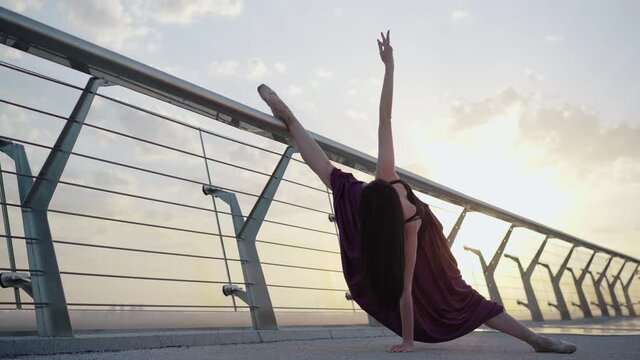 Professional ballerina standing in split on bridge with sun shining at the background. Wide shot portrait of Caucasian elegant ballet dancer moving at sunrise outdoors.