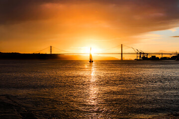 sunset, sailboat stands in front of the sun, reflection on the sea, bridge and buildings behind