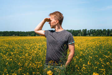Fototapeta premium European traveler man near a yellow flower in a field of rapeseed Sunny day. Happy person outdoors next to plants in full bloom. View of the countryside from a field of rapeseed growing oilseeds