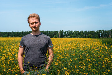Naklejka na ściany i meble European traveler man near a yellow flower in a field of rapeseed Sunny day. Happy person outdoors next to plants in full bloom. View of the countryside from a field of rapeseed growing oilseeds