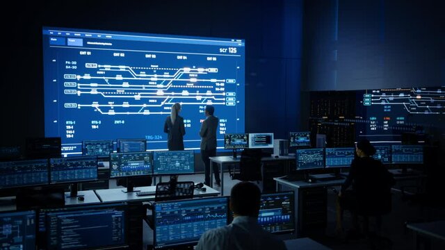 Project Manager and Computer Science Engineer Talking while Using Big Screen Display Showing Infrastructure Infographics and Data.Telecommunications Company System Control and Monitoring Room