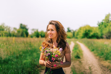Naklejka na ściany i meble Portrait of a beautiful girl with long hair on a country road in summer nature holding a bouquet of colorful flowers, a banner for beauty products or ecology, a place for text