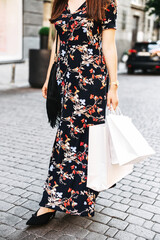 Several paper shopping bags in female hands outdoors. A woman in stylish long summer dress walks...