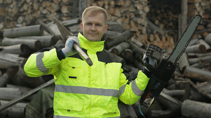 Lumberjack worker. Man woodcutter holds big axe and electric chainsaw in his hands. Firewood
