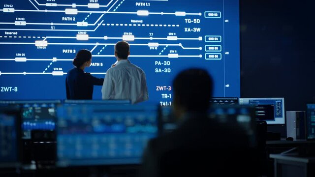 Project Manager and Computer Science Engineer Talking while Using Big Screen Display Showing Infrastructure Infographics and Data.Telecommunications Control Monitoring Room with People Working