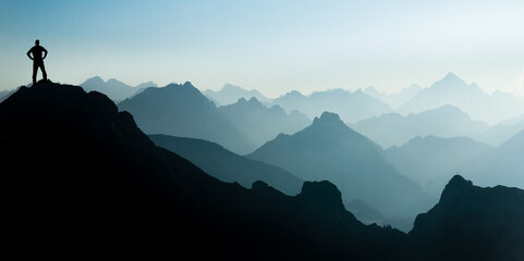 Man reaching summit after climbing and hiking enjoying freedom and looking towards mountains silhouettes panorama during sunrise. - 363511426