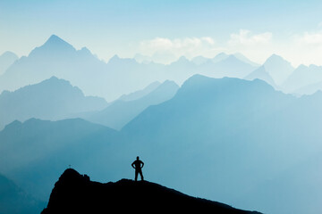 Man reaching summit after climbing and hiking enjoying freedom and looking towards mountains silhouettes panorama during sunrise.