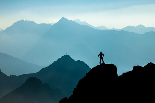 Man reaching summit after climbing and hiking enjoying freedom and looking towards mountains silhouettes panorama during sunrise.