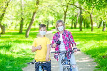 Mother and her young son wearing medical protective mask ride bikes in a summer park and shows thumbs up gesture