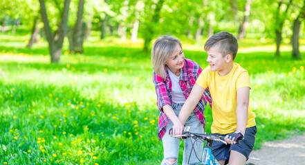 Fototapeta premium Happy family. Mom talks with her young son who ride a bike in summer park. Empty space for text