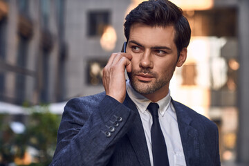 confident young man in full suit talking on the mobile phone and looking away while standing outdoors with cityscape in the background.