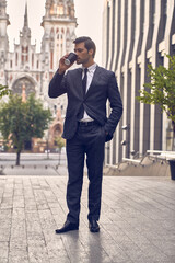 Coffee break. Confident young man in full suit holding coffee cup and looking away while standing outdoors with cityscape in the background.