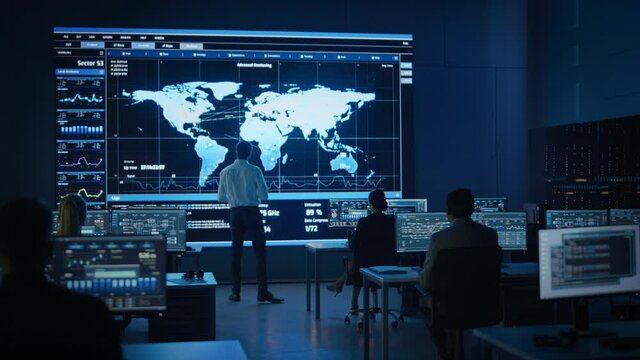 Project Manager Uses Digital Tablet Looks at Big Screen Display Showing Infographics and Global World Map Data.Telecommunications Control Monitoring Room with People Working