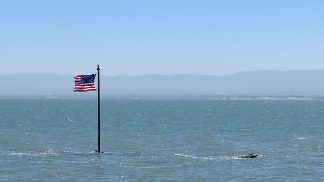 Stars and stripes blowing in the wind on flag pole coming out the ocean with waves and mountains on the horizon