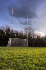 Green fresh grass in focus. Irish National sport goal posts out of focus.Concept practice rugby, hurling, camogie, gaelic footbal. Blue cloudy sky. Vertical image. - obrazy, fototapety, plakaty