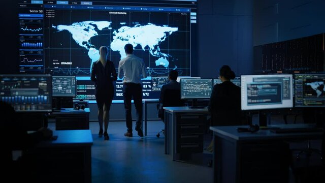 Project Manager Uses Digital Tablet Talks with Computer Scientist Looks at Big Screen Display Showing Infographics Global World Map Data.Telecommunications Control Control Room with People Working