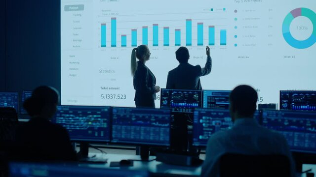 Project Leader, Chief Executive Discuss Data Shown on Big Display. Screens Show Infographics, Charts, Finance Analysis, Stock Market, Growth.Telecommunications Control Room with Working Professionals