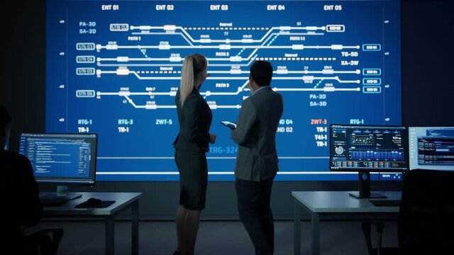 Project Manager and Computer Science Engineer Talking while Using Big Screen Display Showing Infrastructure Infographics and Data. Telecommunications Company System Control Monitoring Room. Zoom out