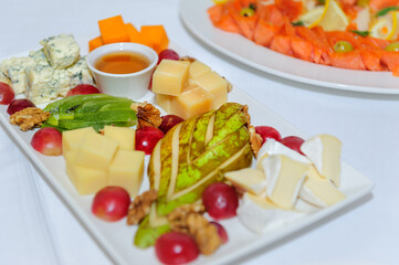 Cheese plate at banquet, close up. Assorted different types of cheeses with honey decorated with nuts, pear, kiwi, grape on large white dish.