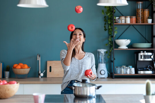 Funny young woman juggling with three red apples in the kitchen at home.