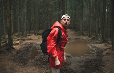 Portrait of a male hiker in a red jacket feeling calm and relaxed in the fir forest. Young tourist man backpacker with serious facial expression is in the mountain woods, puddle on the background.