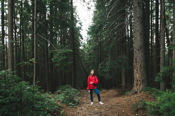 Obraz premium Minimalistic full-length photo of hiker girl in a red jacket in the mountain fir forest. A female traveler wearing a red raincoat is among the trees in the woods. Concept shot, copy space.