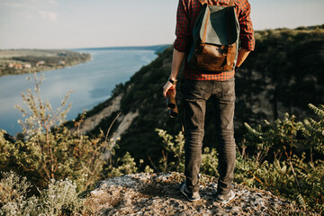 Young hipster man with a backpack, holding binoculars, standing on a cliff in mountains, looking towards a lake. 