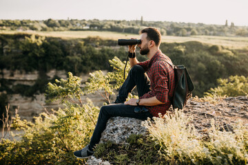 Naklejka na ściany i meble Young hipster man looking through binoculars, sitting on a cliff in mountains, watching landscapes. Concept of tourism.