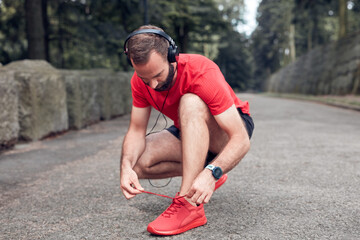 Sporty adult man making pause after jogging and exercising in the park and tying running shoes.